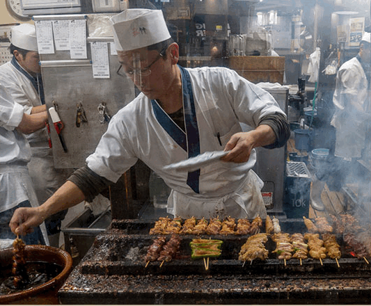A cook at Toritake turning rows of skewers over a long charcoal grill behind the counter